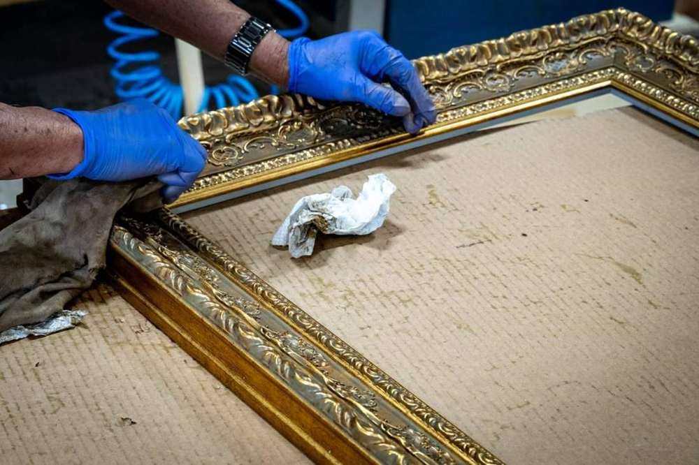 Frame-shop artisan in blue gloves hand-finishing an ornate gold moulding on the Fort Lauderdale production floor